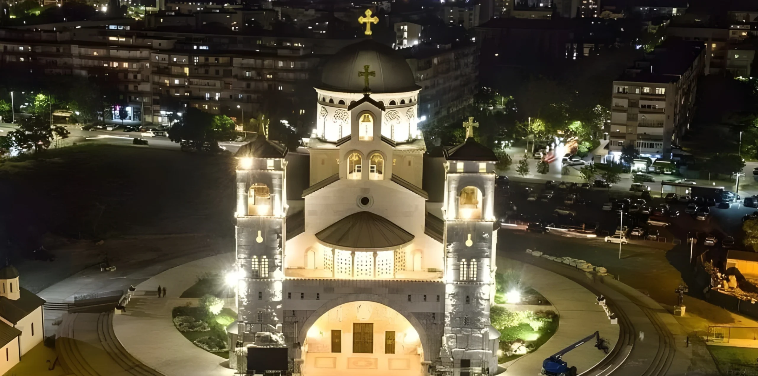 Aerial night view of a brightly illuminated cathedral with central dome and twin towers, surrounded by city streets, parked cars, and residential buildings.