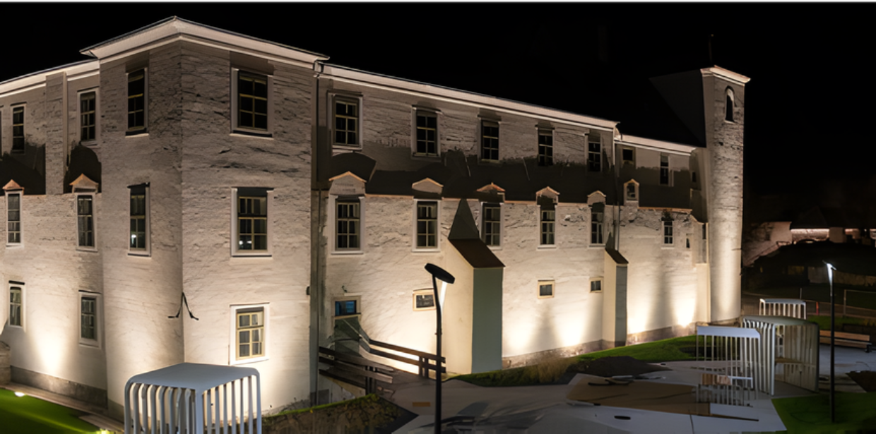 Historic stone building illuminated at night with exterior architectural lighting highlighting windows, walls, and a tower structure in a landscaped setting.