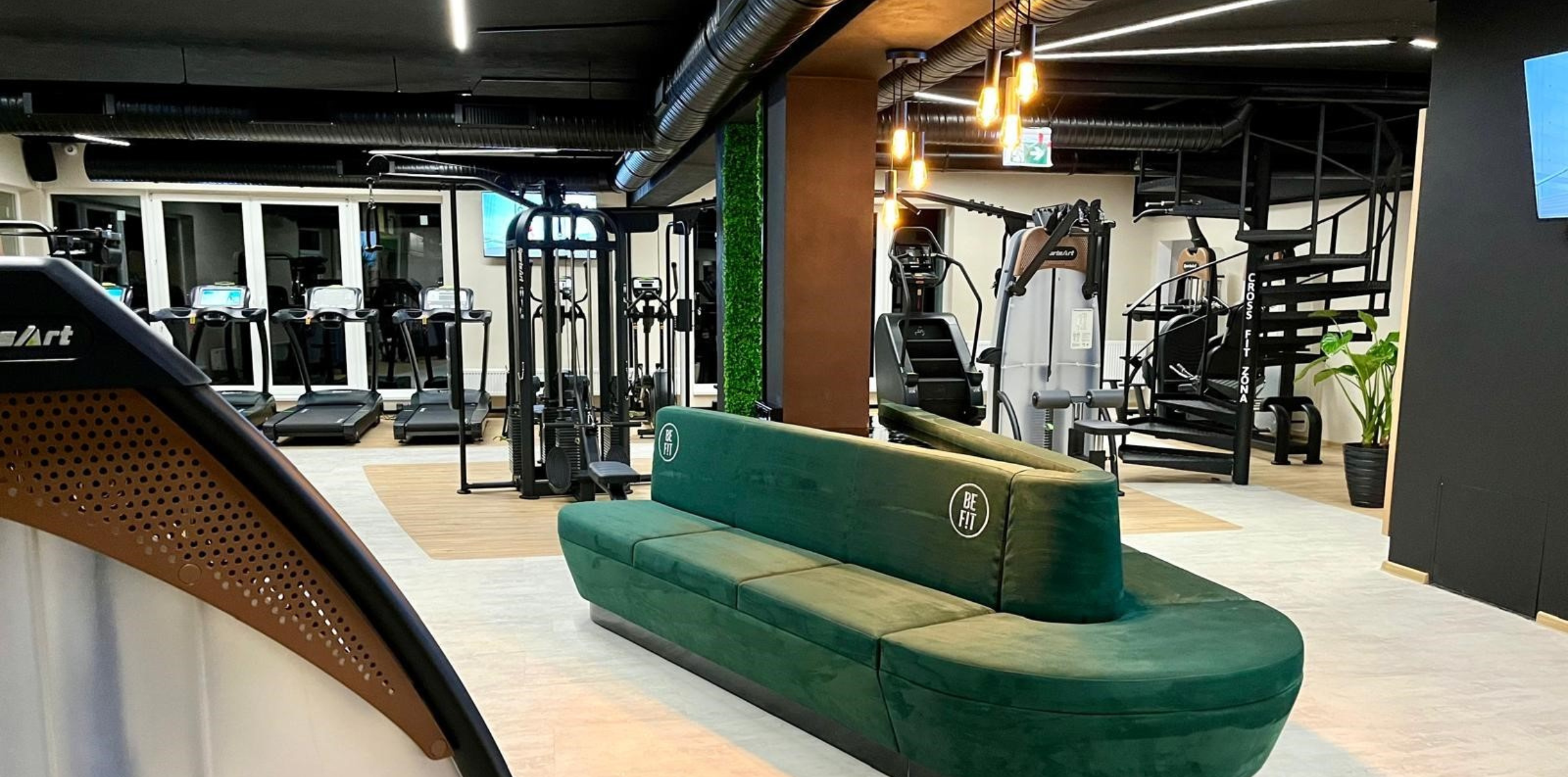 Modern gym interior featuring workout machines, treadmills, a green upholstered seating unit, and exposed ceiling ductwork with warm pendant lighting.