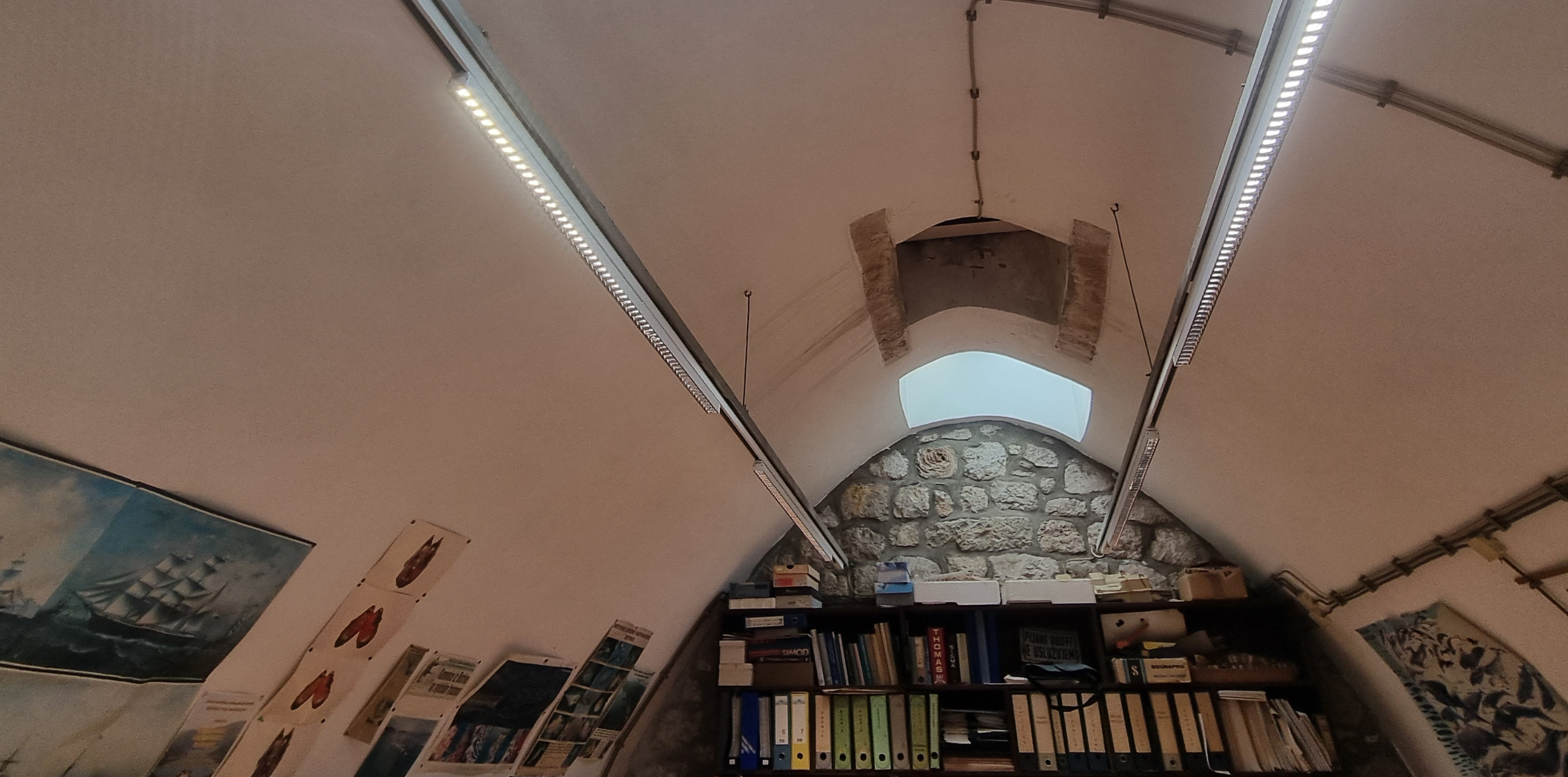Interior view of a room with a curved, vaulted ceiling lit by two long linear LED fixtures suspended on wires. The lights run parallel along the length of the ceiling. At the far end is a stone wall with a small arched window above a shelf filled with books, binders, and miscellaneous items. Framed pictures and posters are mounted on the side walls, and cable conduits run along the ceiling edges.